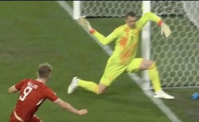 A soccer goalkeeper in a bright yellow uniform dives dramatically to his right, arms outstretched, while a player in a red…