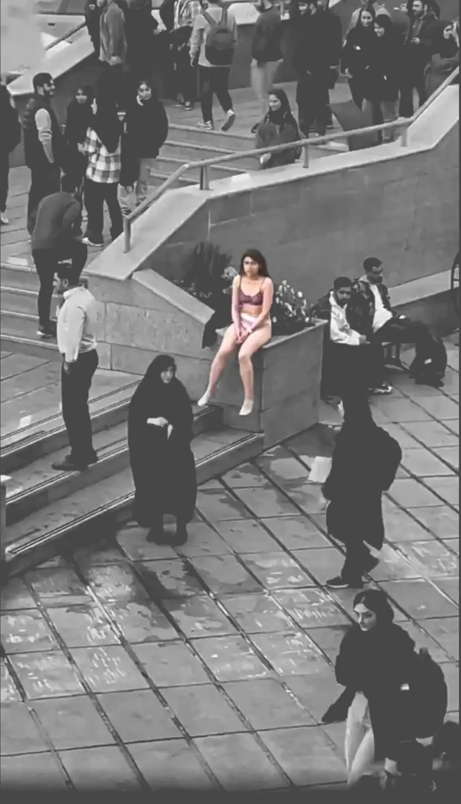 A black-and-white surveillance-style photo shows a young woman in a bikini top and shorts sitting on a concrete ledge in a…
