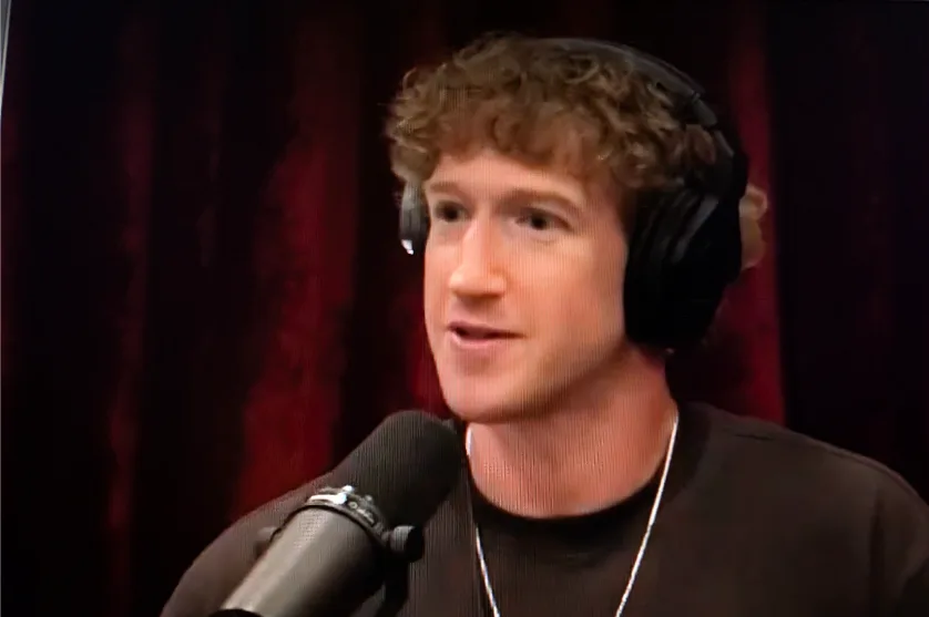 a man with curly brown hair wearing black headphones and a black shirt speaks into a microphone against a red curtain…