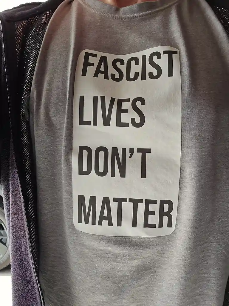 A close-up photo shows a person wearing a gray t-shirt with a white rectangular patch that reads 'FASCIST LIVES DON'T…