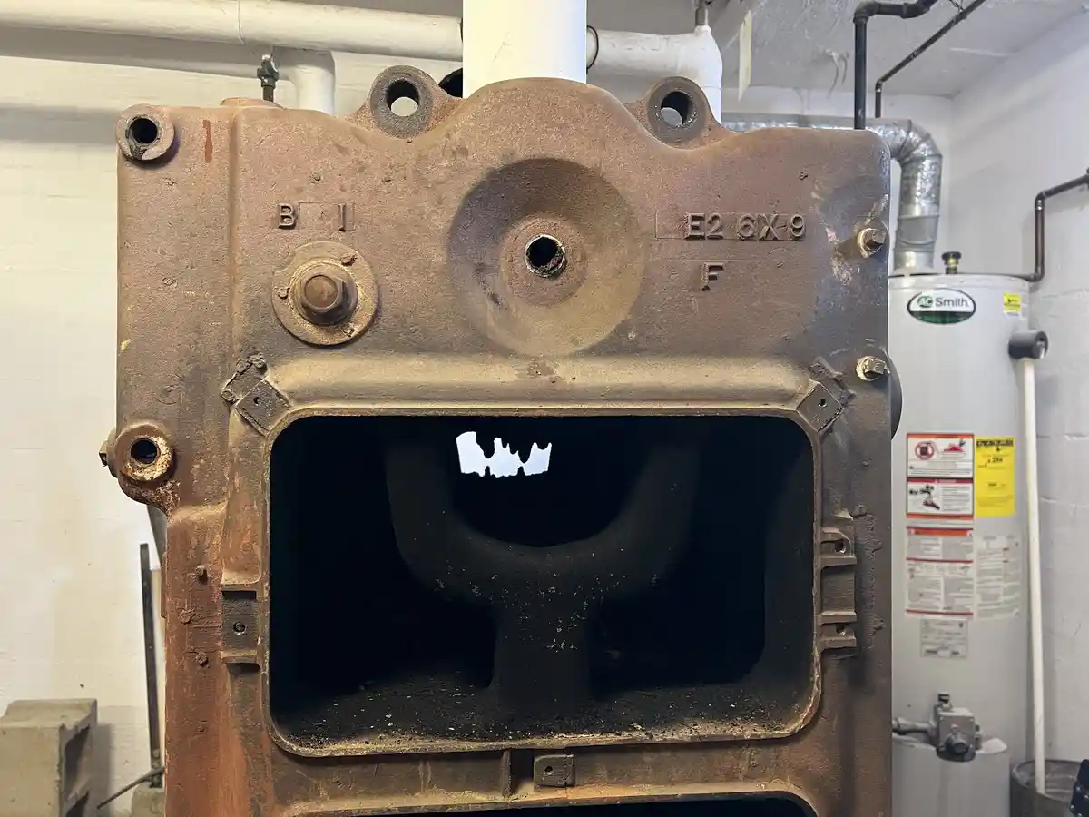 A rusty, industrial metal boiler or furnace with a large rectangular opening. Inside the opening, a dark, curved pipe is…