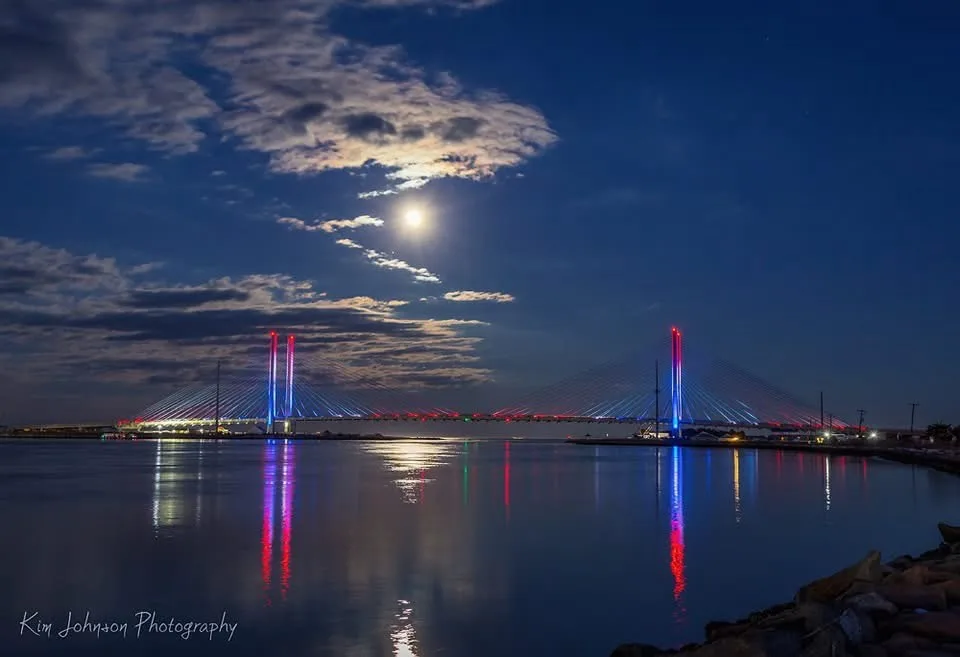 A serene night photograph of a modern cable-stayed bridge illuminated with red and blue lights, its reflection shimmering on…