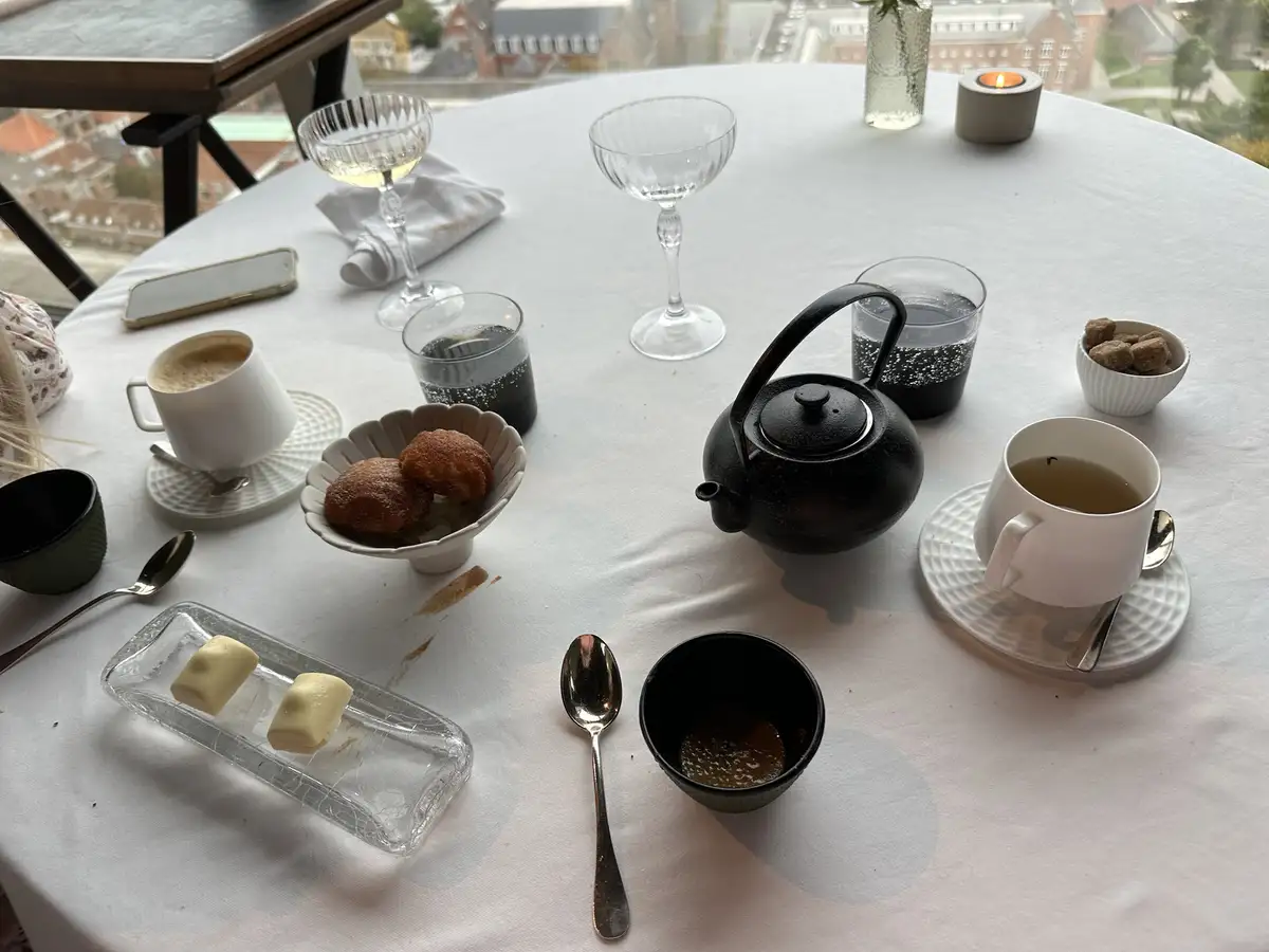 A round table set for tea with a black teapot, white teacups, glasses, pastries, and a candle, with visible crumbs and…