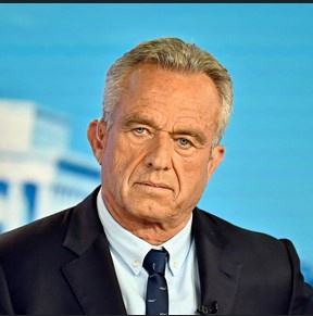 A close-up photo of a middle-aged man with graying hair, wearing a dark suit and tie, looking sternly at the camera. Behind…