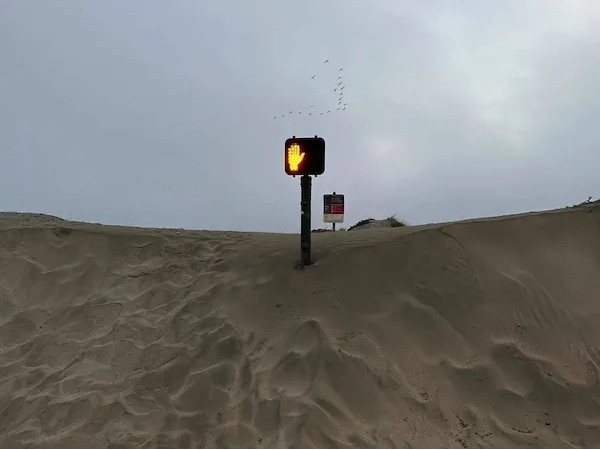A pedestrian crossing signal displaying a glowing yellow hand is mounted on a pole atop a large sand dune under an overcast…