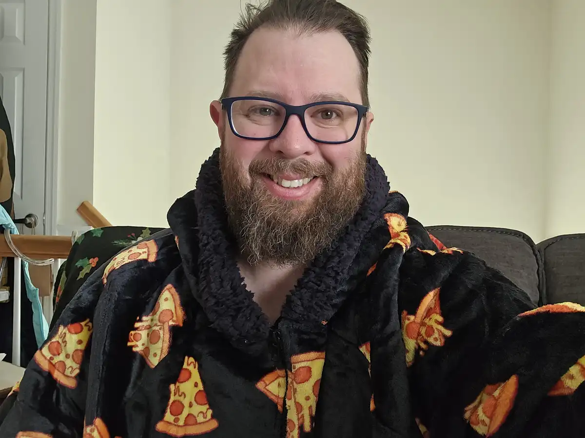 A smiling man with glasses and a beard wears a black fleece robe covered in pizza slice patterns while sitting on a couch…