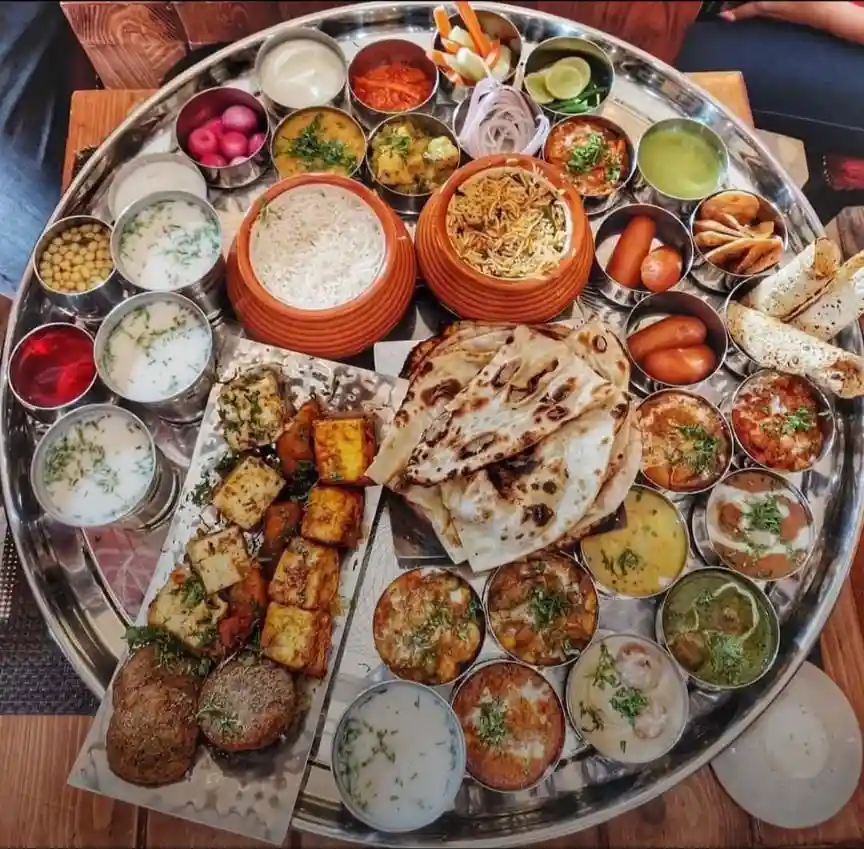a large, round metal platter holds an extensive indian meal called a thali, featuring multiple small bowls and plates with…