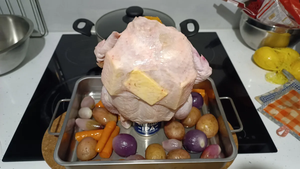 A raw, whole chicken sits upright in a roasting pan filled with vegetables, with a large pat of butter on its back. The…