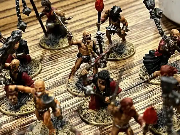 A close-up photo shows a collection of painted miniature orc figurines arranged on a wooden surface, each holding weapons…