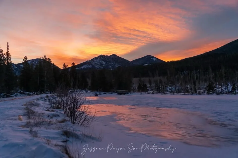 A serene winter landscape photograph showing snow-covered ground, a frozen lake reflecting a vibrant orange and pink sunset…