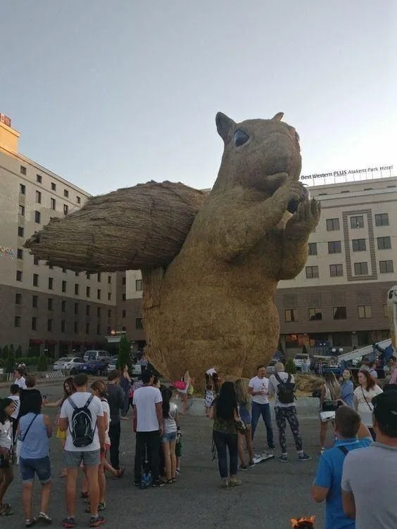 A giant, hay-like sculpture of a squirrel holding a nut stands in a city plaza, surrounded by people observing it. The…