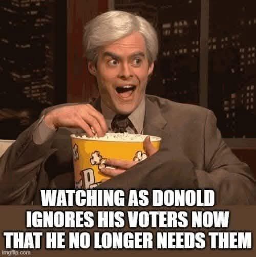 A man with gray hair and a suit sits on a couch holding a large yellow popcorn bucket, looking shocked with wide eyes and an…