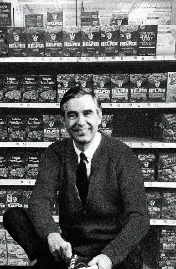 a black-and-white photo shows a smiling man in a sweater and tie sitting on a supermarket floor surrounded by shelves…
