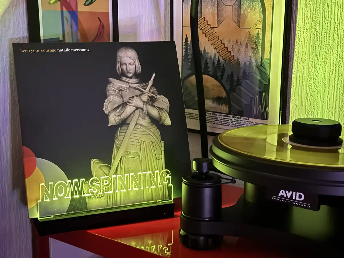 A vinyl record player with a glowing neon sign that reads 'NOW SPINNING' sits next to the album cover for Natalie Merchant's…
