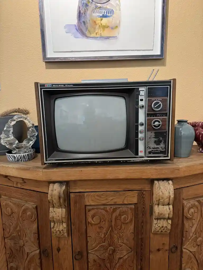 An old Hitachi television with a wooden frame and knobs sits on a carved wooden cabinet, its screen blank and reflecting a…