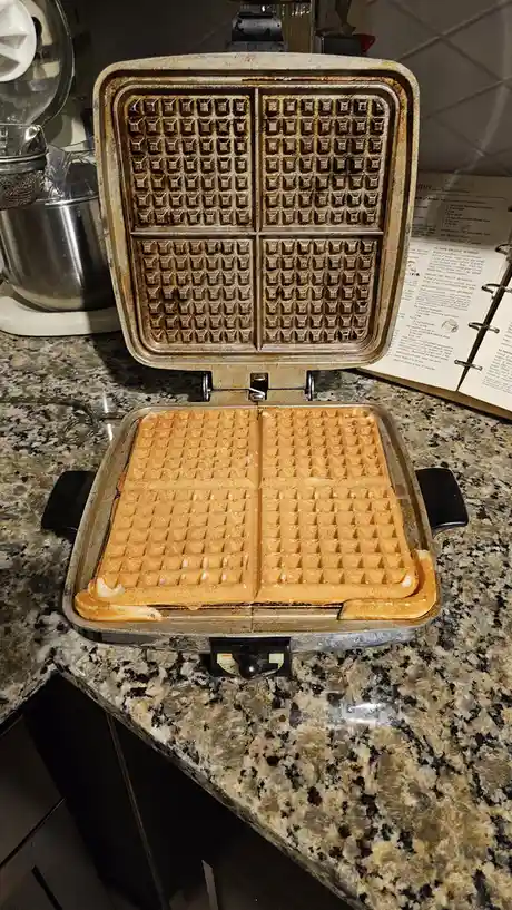 A close-up photo shows an open, well-used waffle maker on a granite countertop, with two golden-brown waffles stuck to the…