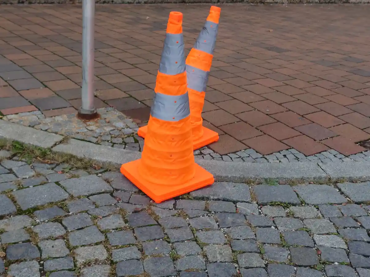 Two orange traffic cones with reflective stripes are standing upright on a paved sidewalk next to a metal pole, with one…
