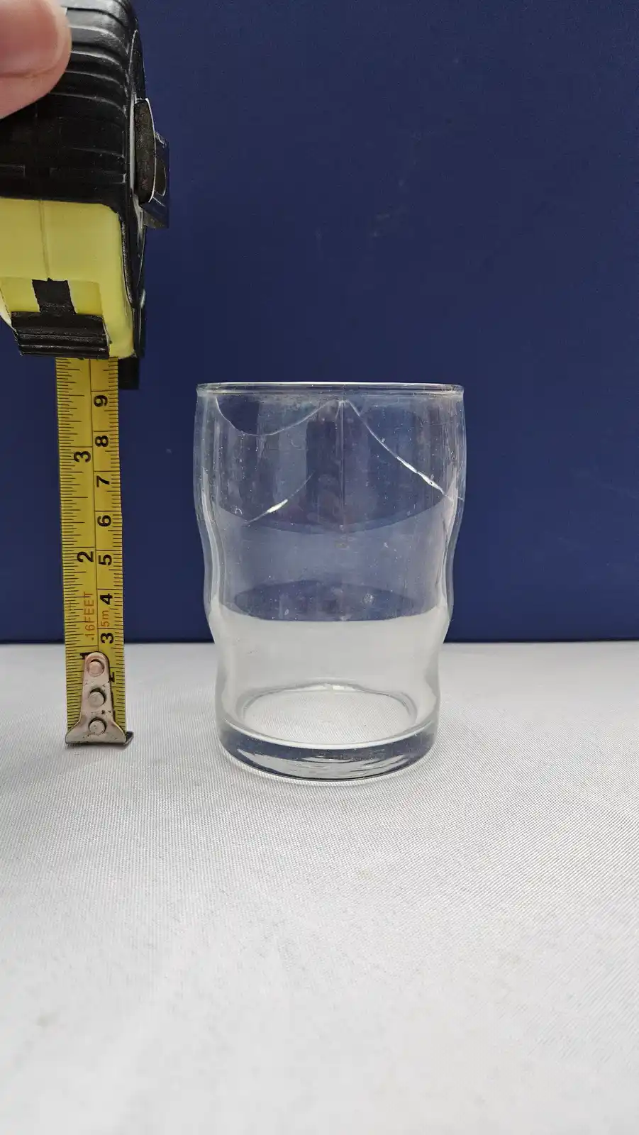 A clear, empty drinking glass sits on a white surface next to a yellow tape measure held by a hand, with the tape extended…