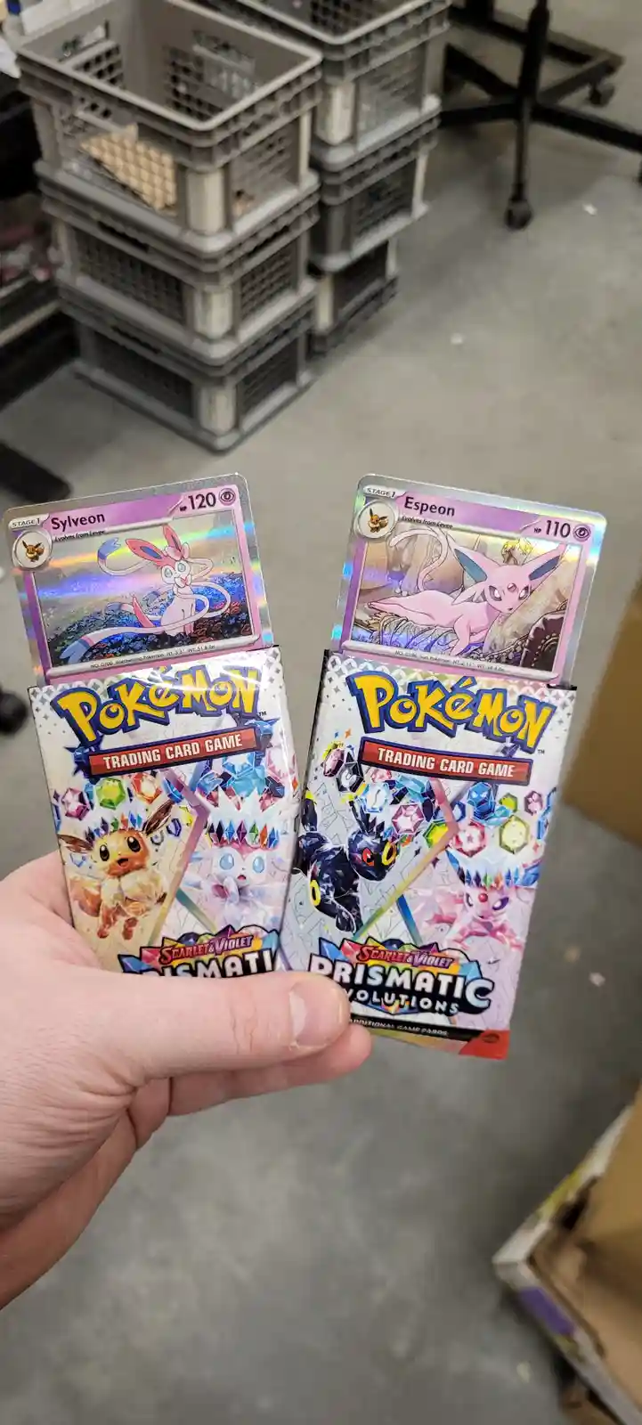 A hand holds two Pokémon Trading Card Game booster packs from the Scarlet & Violet Prismatic Evolutions set, each with a…