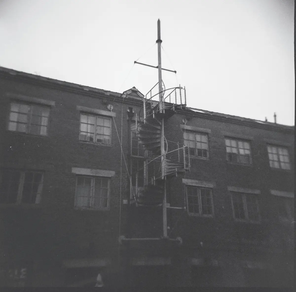 A black-and-white photo shows a brick building with a spiral metal staircase leading up to a rooftop antenna structure. The…