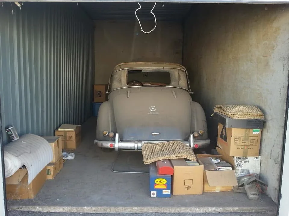 A vintage, dusty, beige convertible car is parked inside a storage container, surrounded by cardboard boxes, a rolled-up…