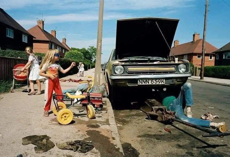 A vintage car with its hood open sits on a residential street, with a person lying underneath it, appearing to work on the…