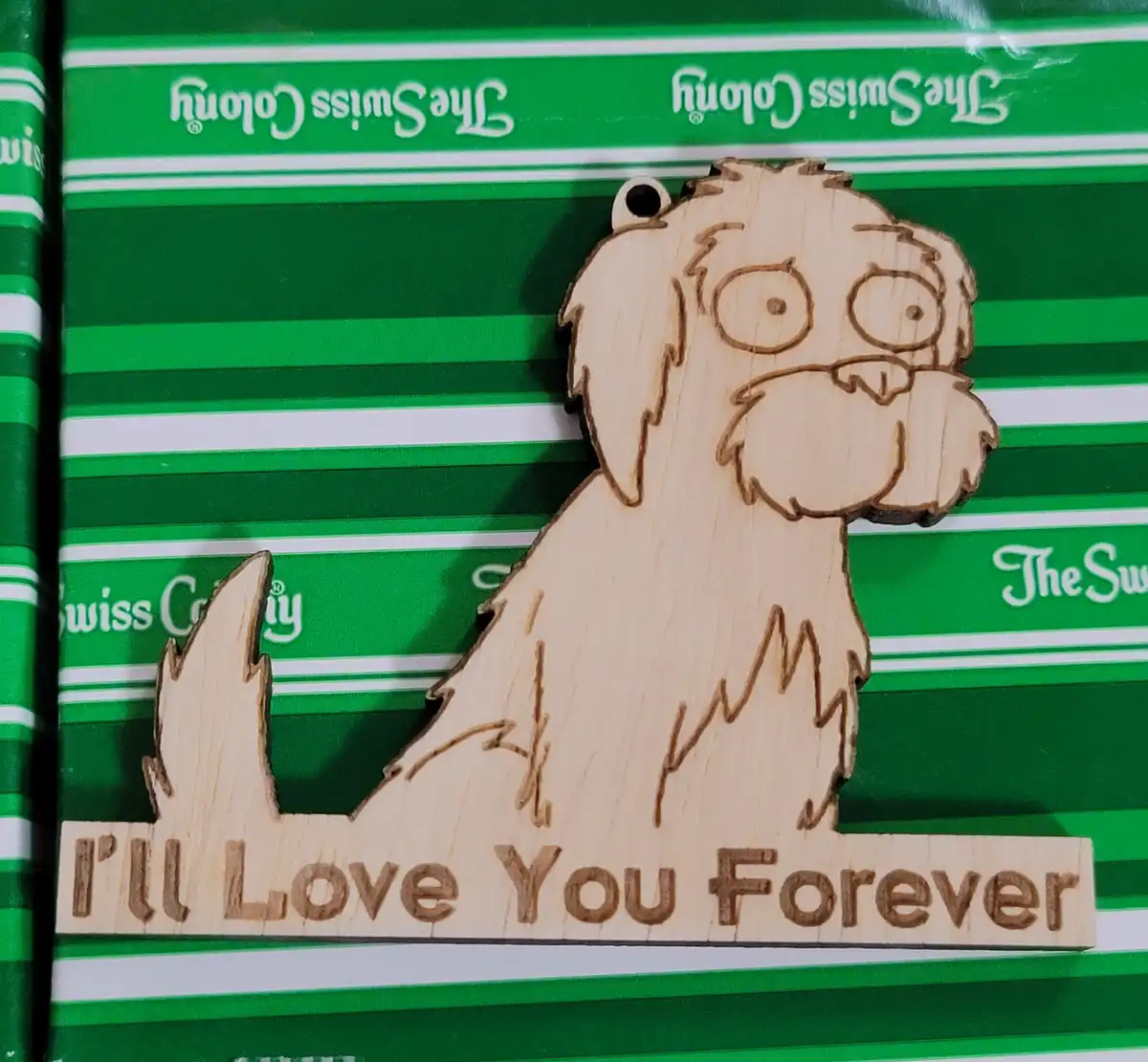 A wooden cutout of a cartoon dog with wide eyes and a mustache sits on green and white striped wrapping paper branded 'The…