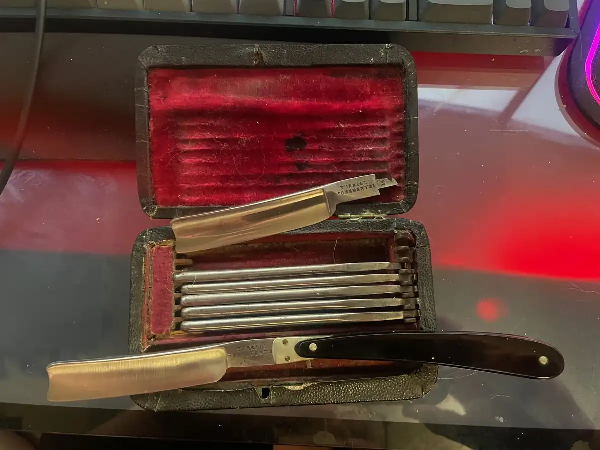 An open, worn black leather case with red velvet lining sits on a desk, containing a straight razor and multiple spare…