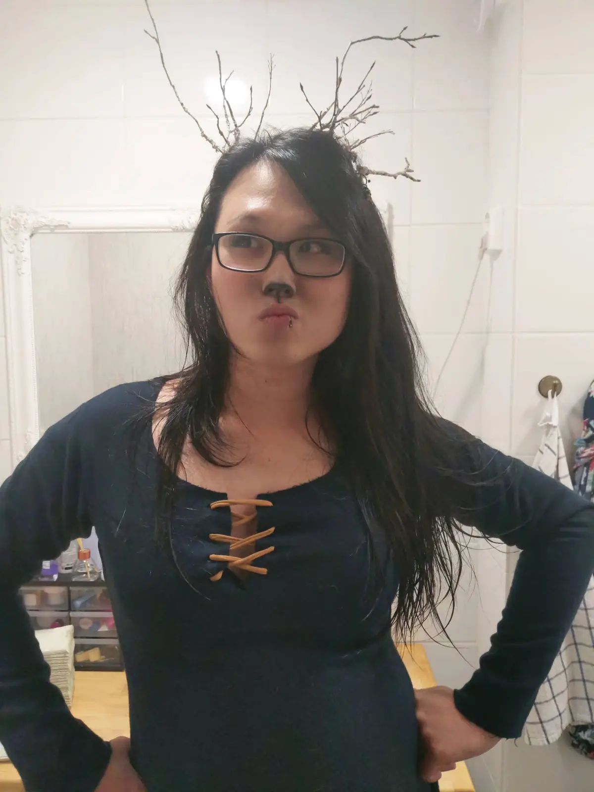 A person with long dark hair and glasses poses in a bathroom, wearing a dark blue laced top and a crown of bare twigs on…