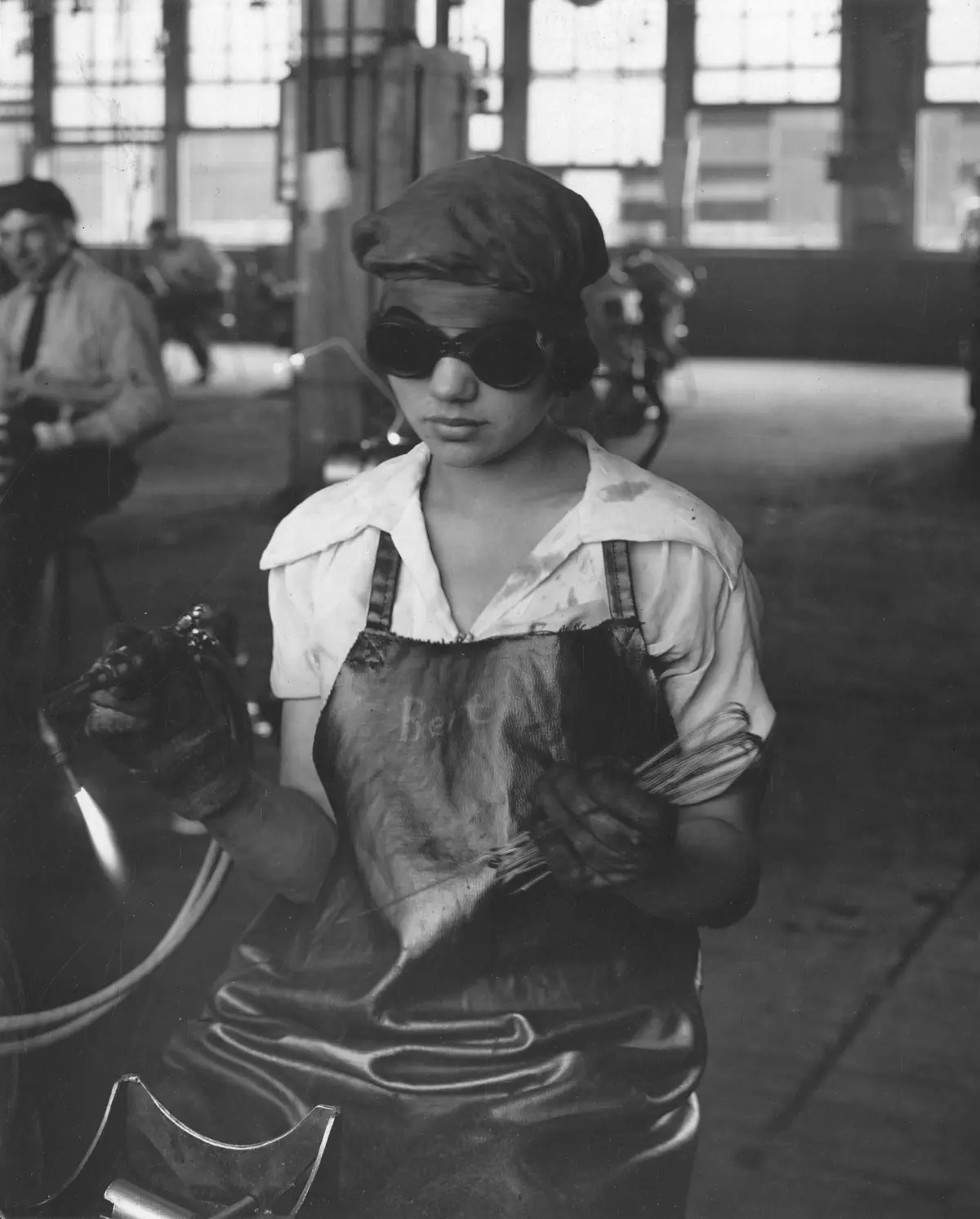 A black-and-white historical photograph shows a woman in a factory wearing protective goggles, a cap, and a stained apron…
