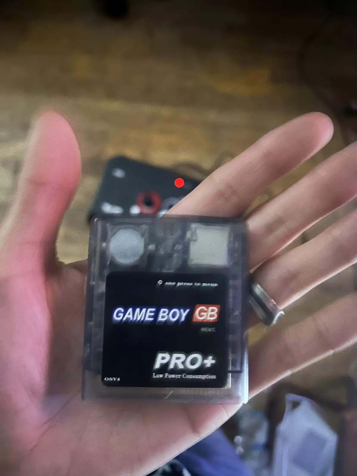 A hand holds a transparent, modified Game Boy Pro+ cartridge with visible internal circuitry. The label reads 'GAME BOY GB…