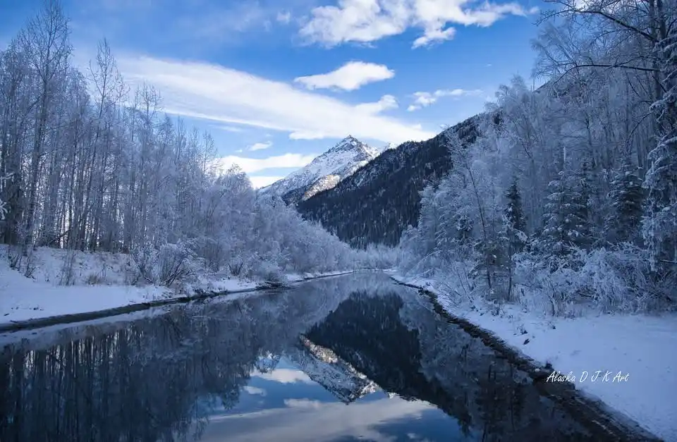 A serene winter landscape photograph shows a snow-covered river flanked by frost-laden trees, reflecting a snow-capped…