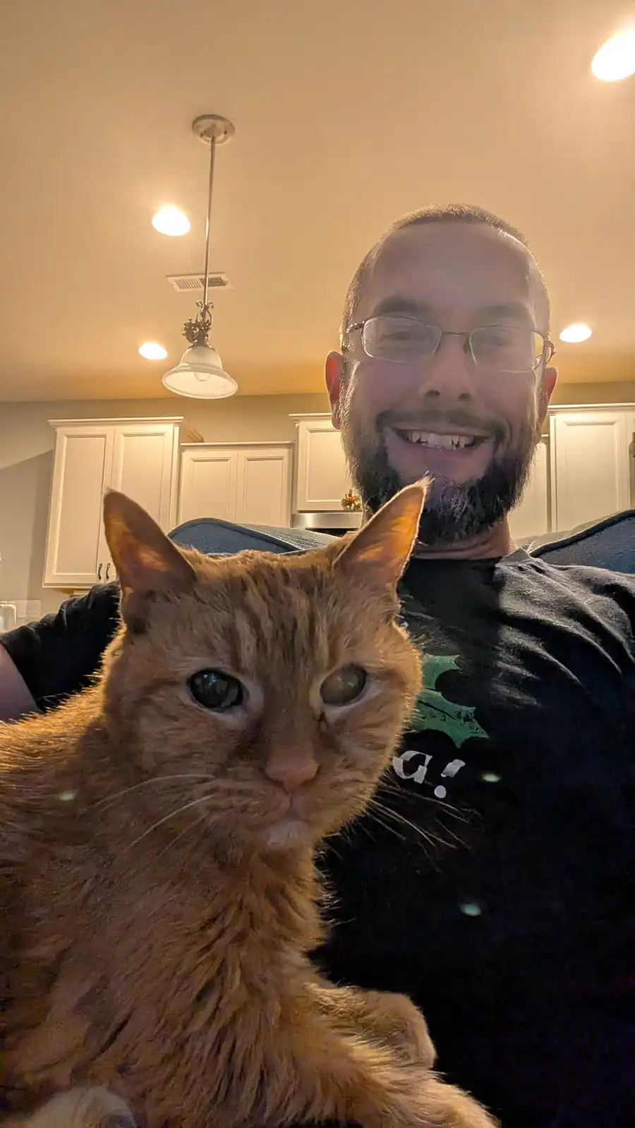 A man with glasses and a beard smiles broadly while holding an orange tabby cat in a selfie taken indoors, likely in a…