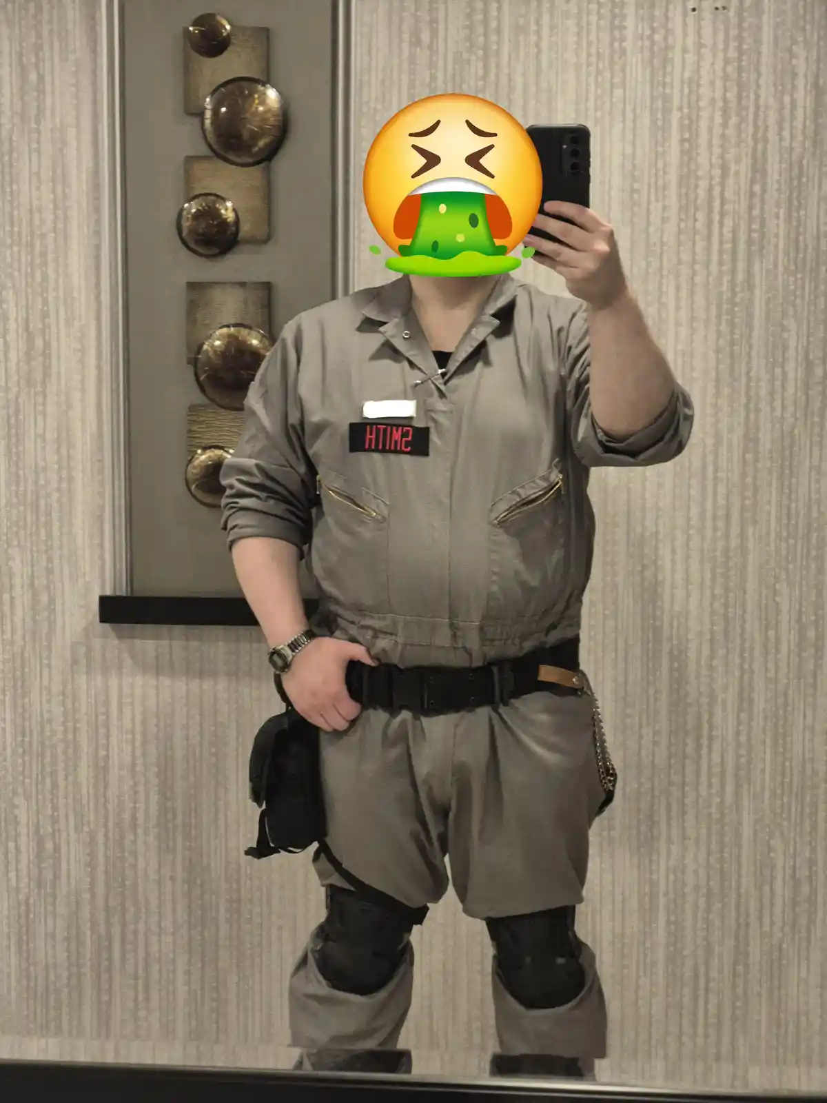 A person in a grey jumpsuit with a name tag reading 'SMITH' and black knee pads takes a mirror selfie while holding a phone…