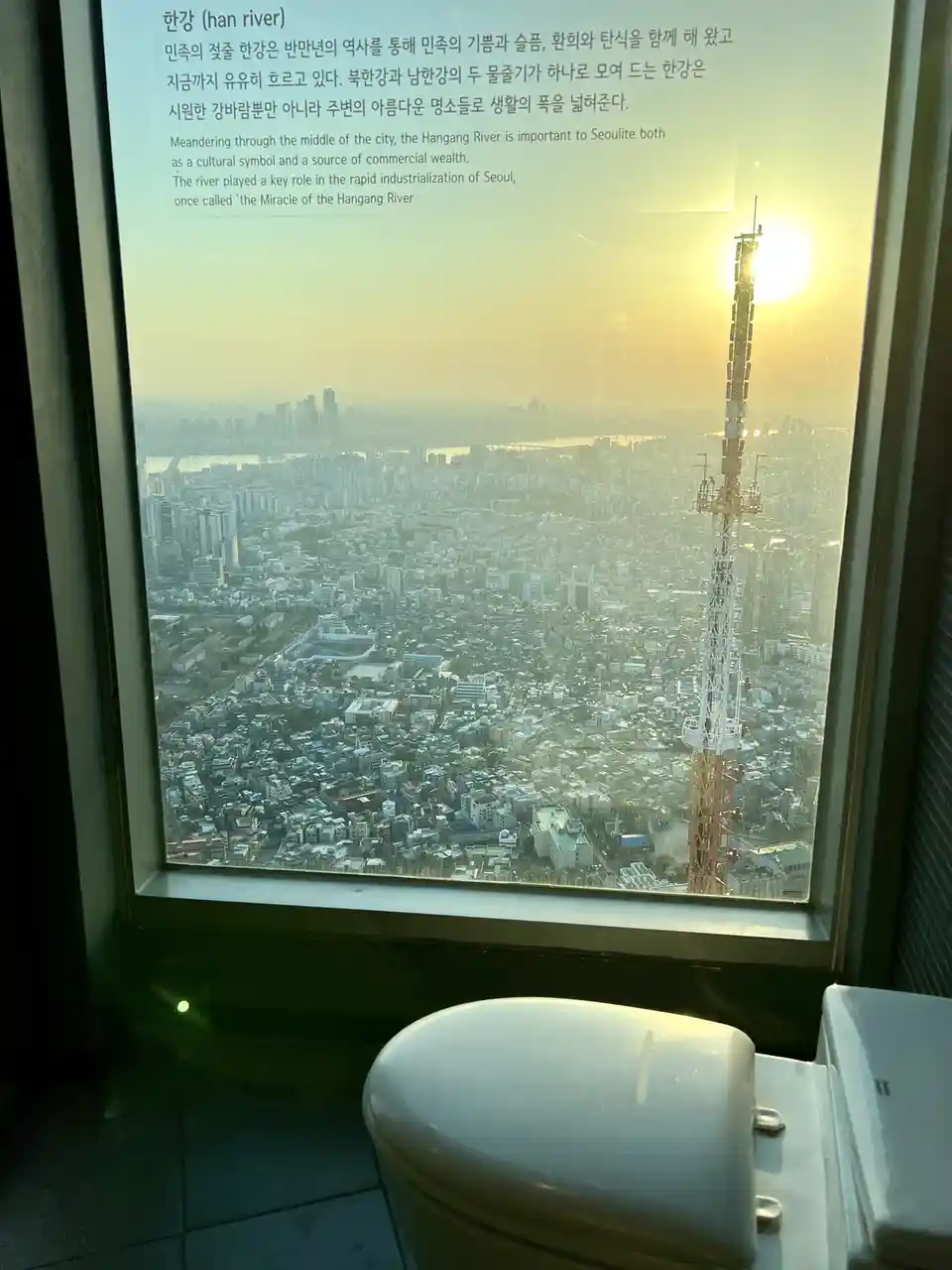 A toilet is visible in the foreground, looking out a window at a sprawling cityscape during sunset, with a tall…