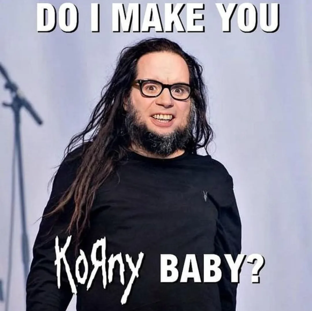 A man with long dark hair, glasses, and a beard wearing a black shirt stands on a stage with a microphone stand visible…