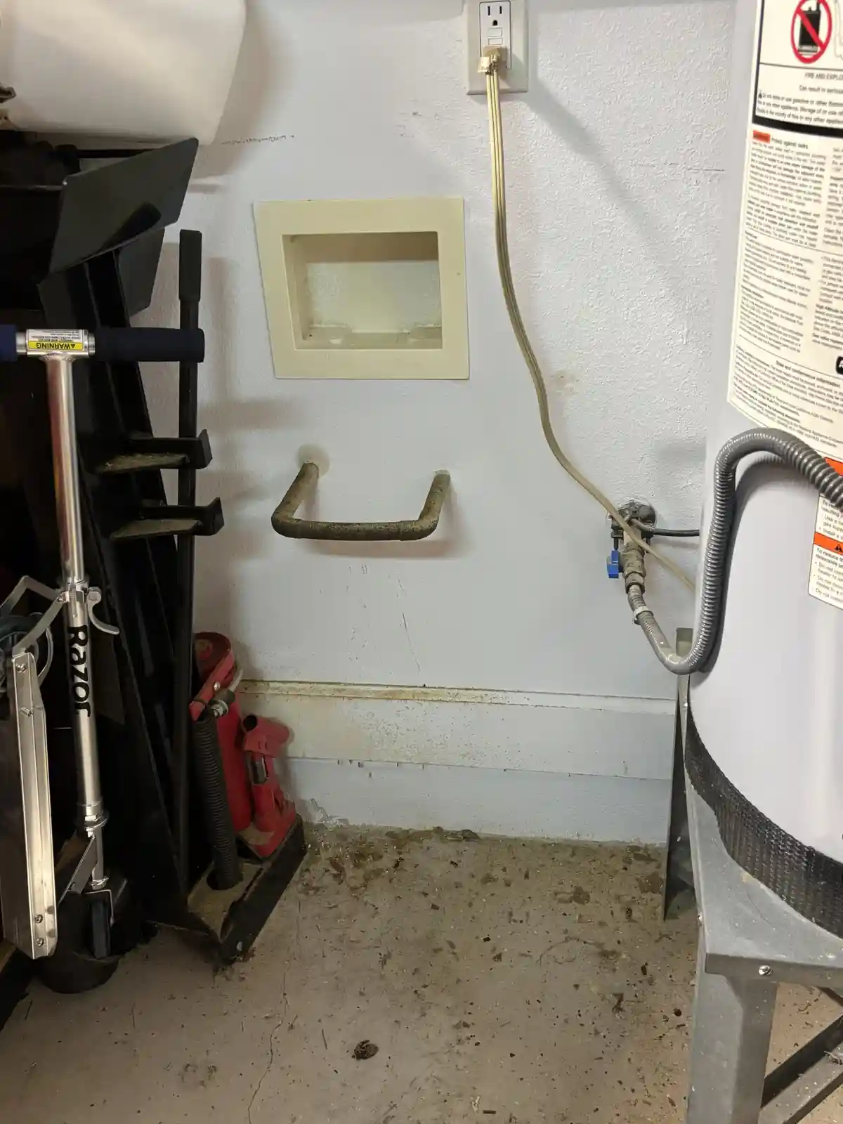A dirty, cluttered utility room corner shows a water heater on the right, a rusty pipe handle on the wall, and a scooter…