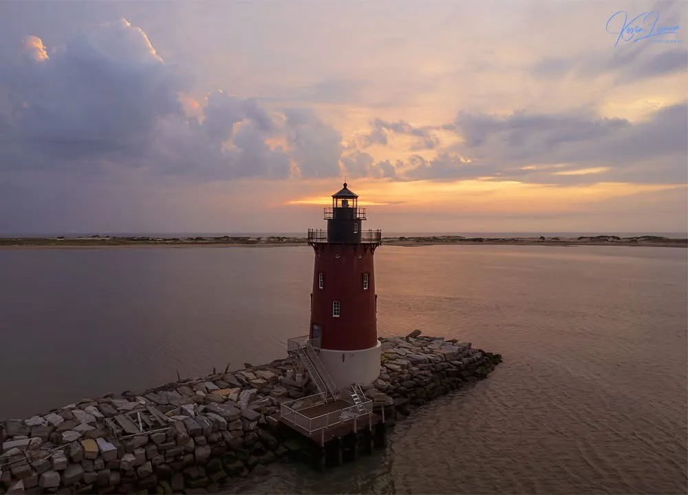 A scenic photo of a red and white lighthouse at sunset, standing on a stone pier extending into calm water under a cloudy…