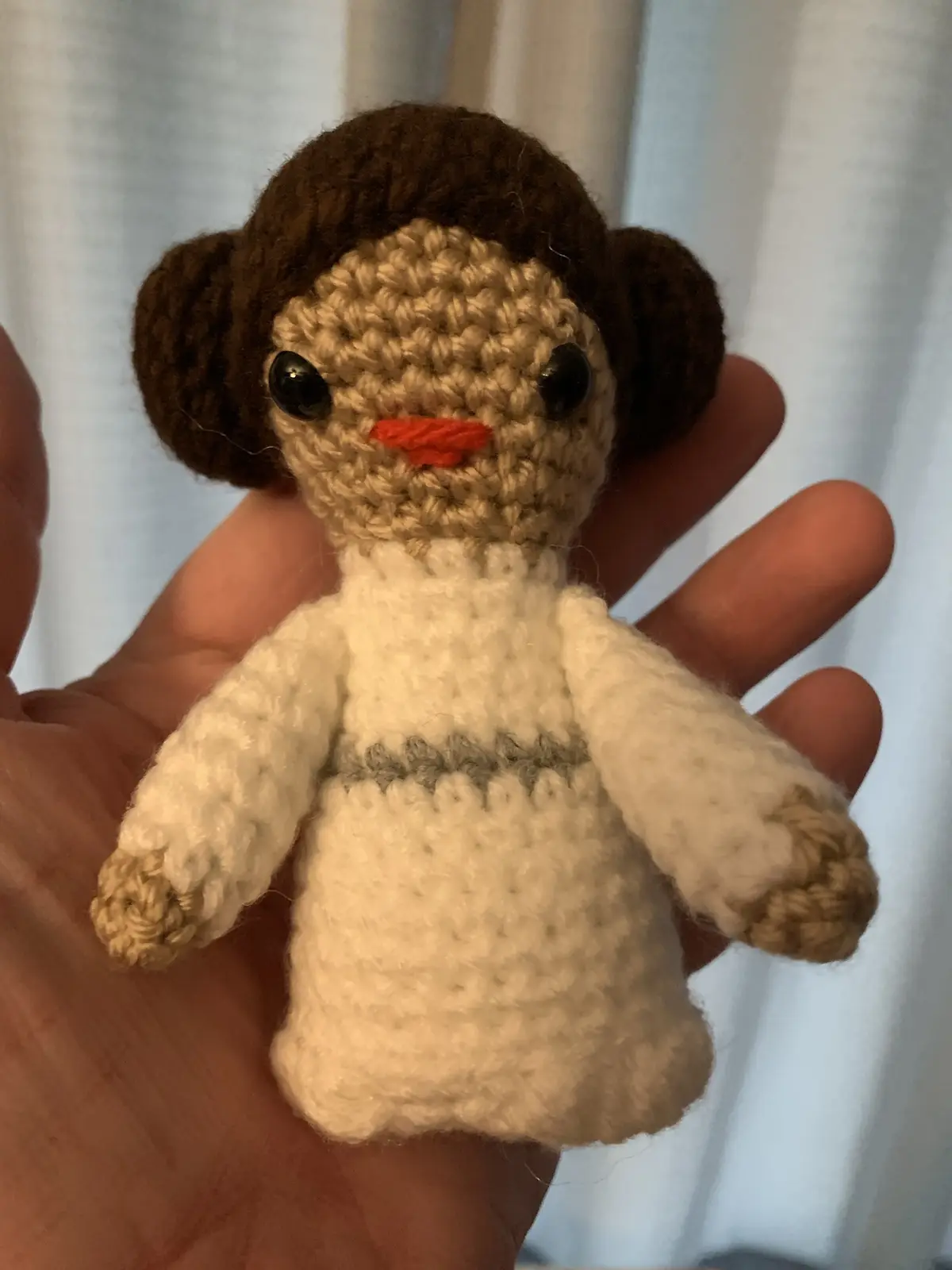 A hand holds a small, crocheted doll resembling Princess Leia from Star Wars, identifiable by her brown buns and white dress…