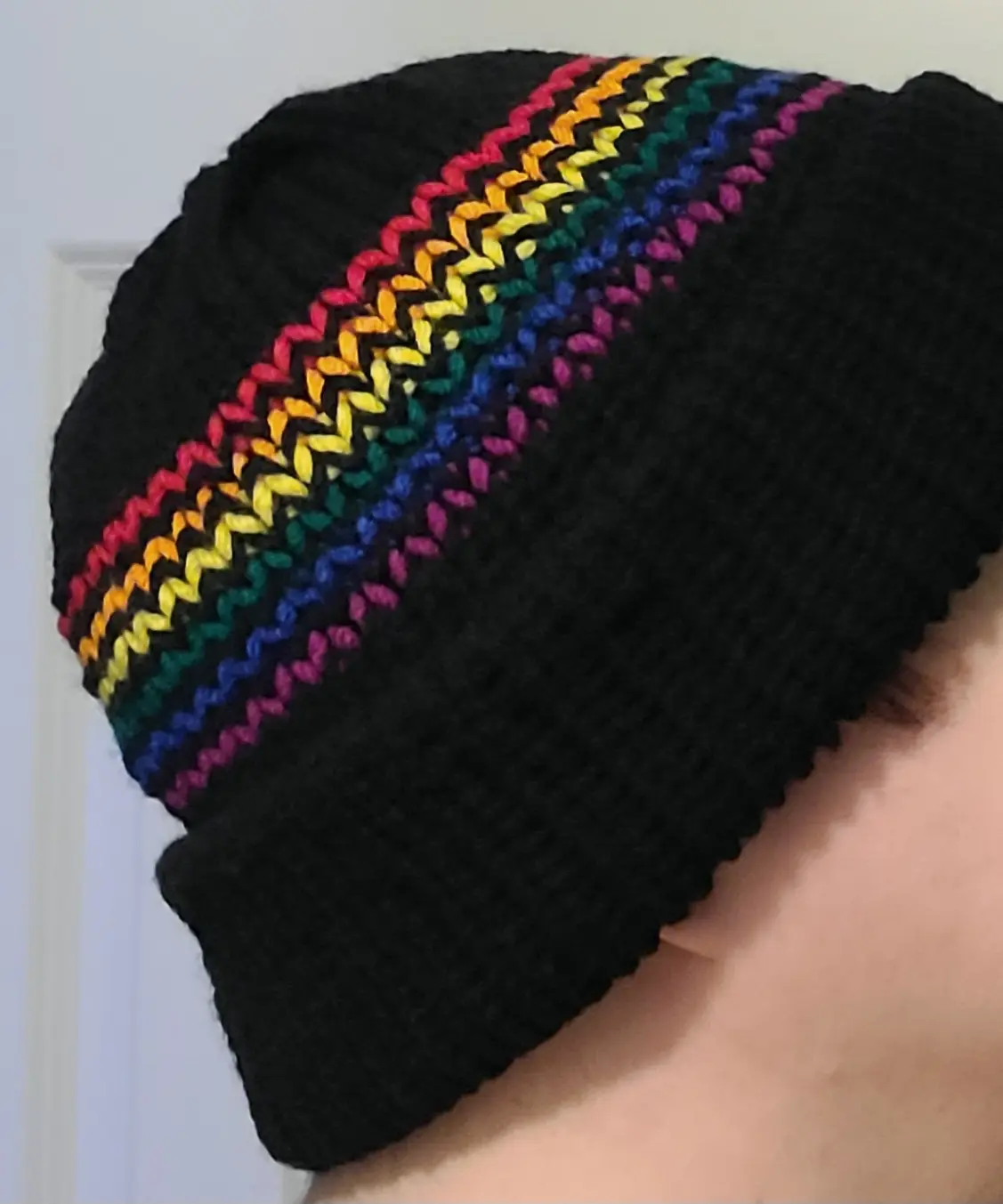 A close-up photo shows the side of a person's head wearing a black knitted beanie with a single, vibrant rainbow-colored…