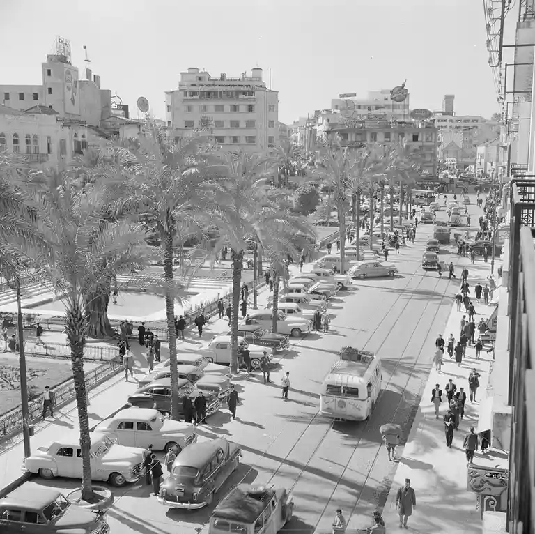a black and white photograph of a bustling city street lined with palm trees and vintage cars from the mid-20th century…