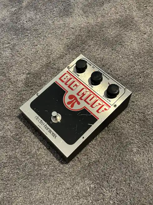 A close-up photo of a vintage Electro-Harmonix Big Muff Pi guitar effects pedal resting on a gray carpet. The pedal has a…