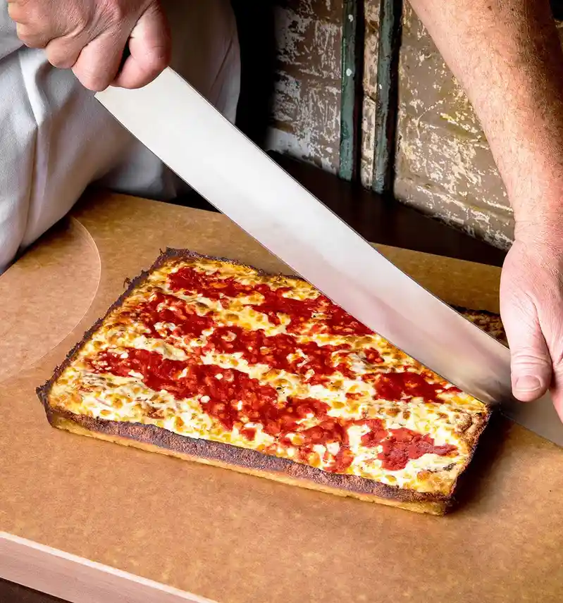 A person in a white chef's coat uses an oversized, long-bladed knife to slice a rectangular, thick-crust pizza with tomato…