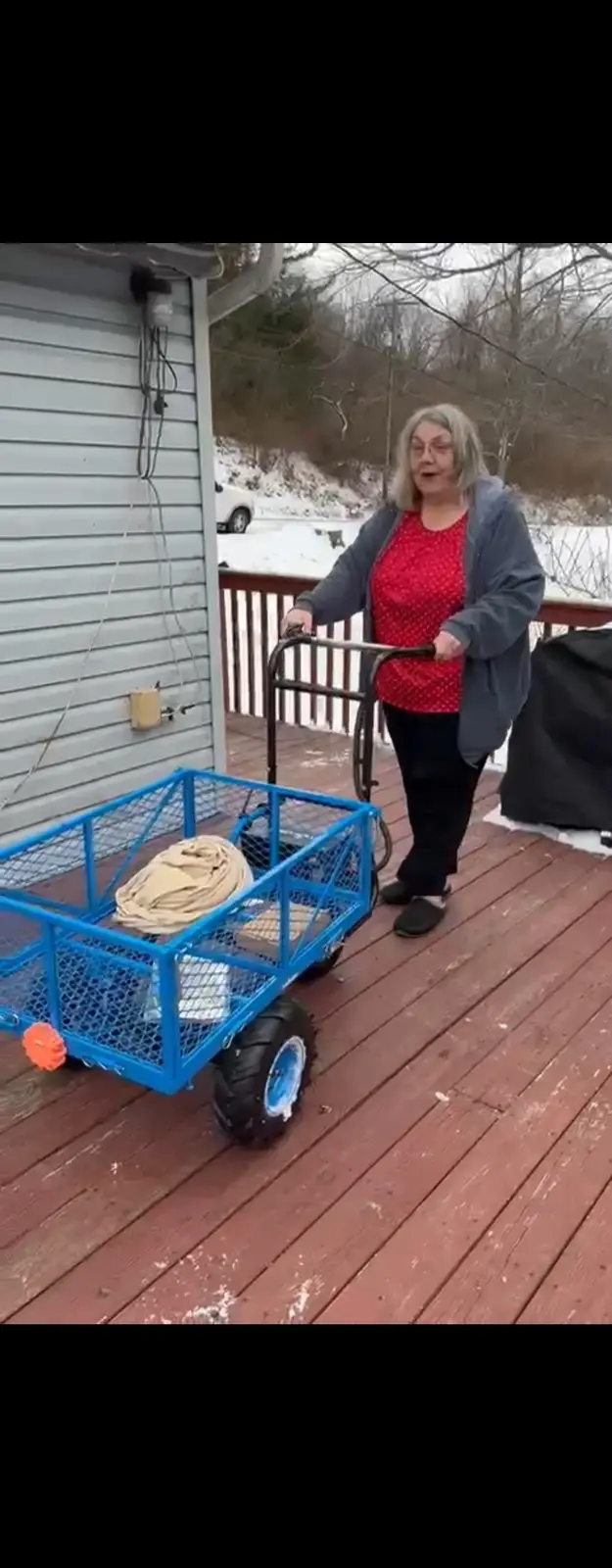 A woman with gray hair and glasses, wearing a red polka-dot shirt and gray jacket, stands on a snowy wooden deck pushing a…