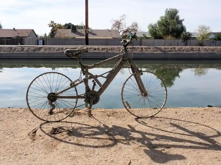 A rusty, abandoned bicycle stands upright on sandy ground beside a calm canal, its frame and wheels covered in dirt and…