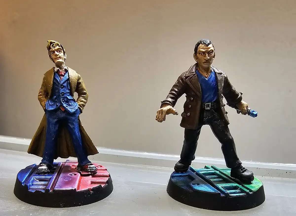 Two hand-painted Doctor Who figurines stand side-by-side on bases; the left figure wears a brown coat over a blue suit with…
