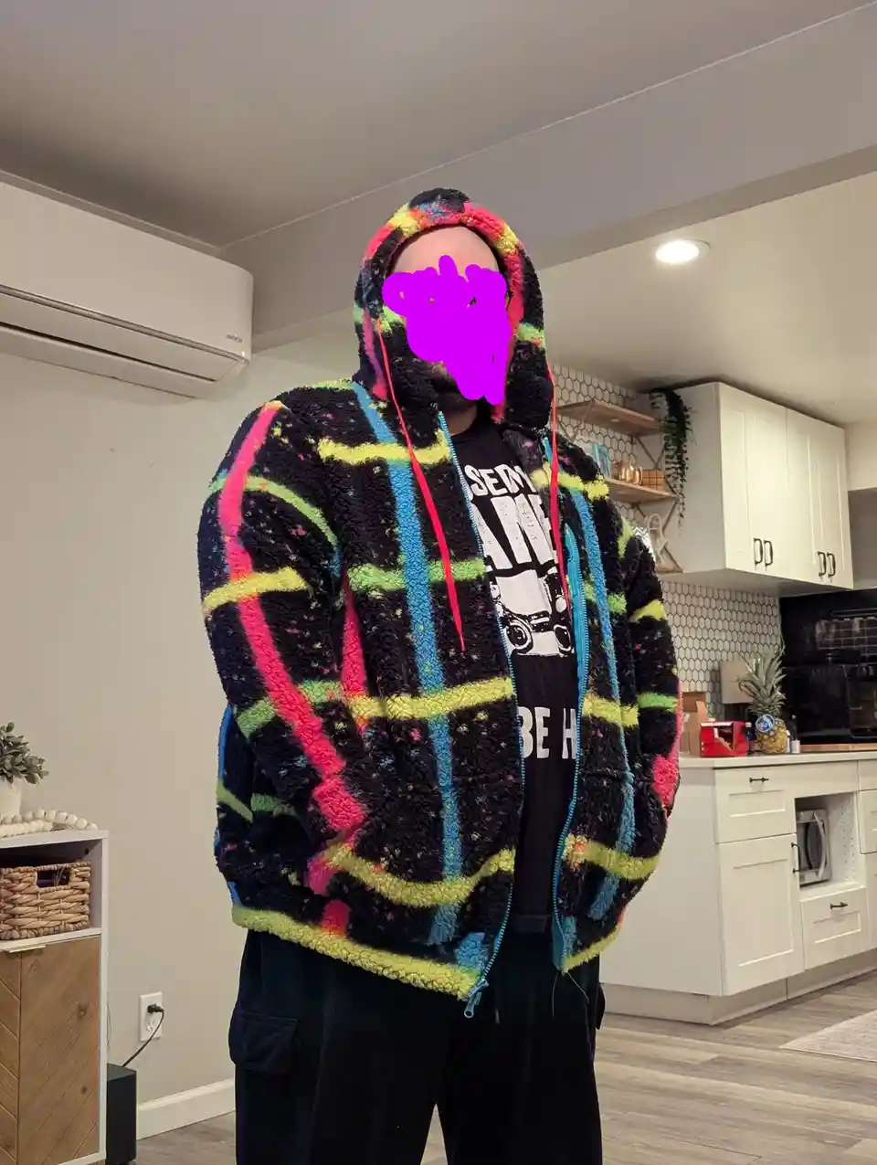 A person stands in a kitchen wearing a very colorful, fuzzy, black hoodie with bright neon stripes in pink, yellow, and…