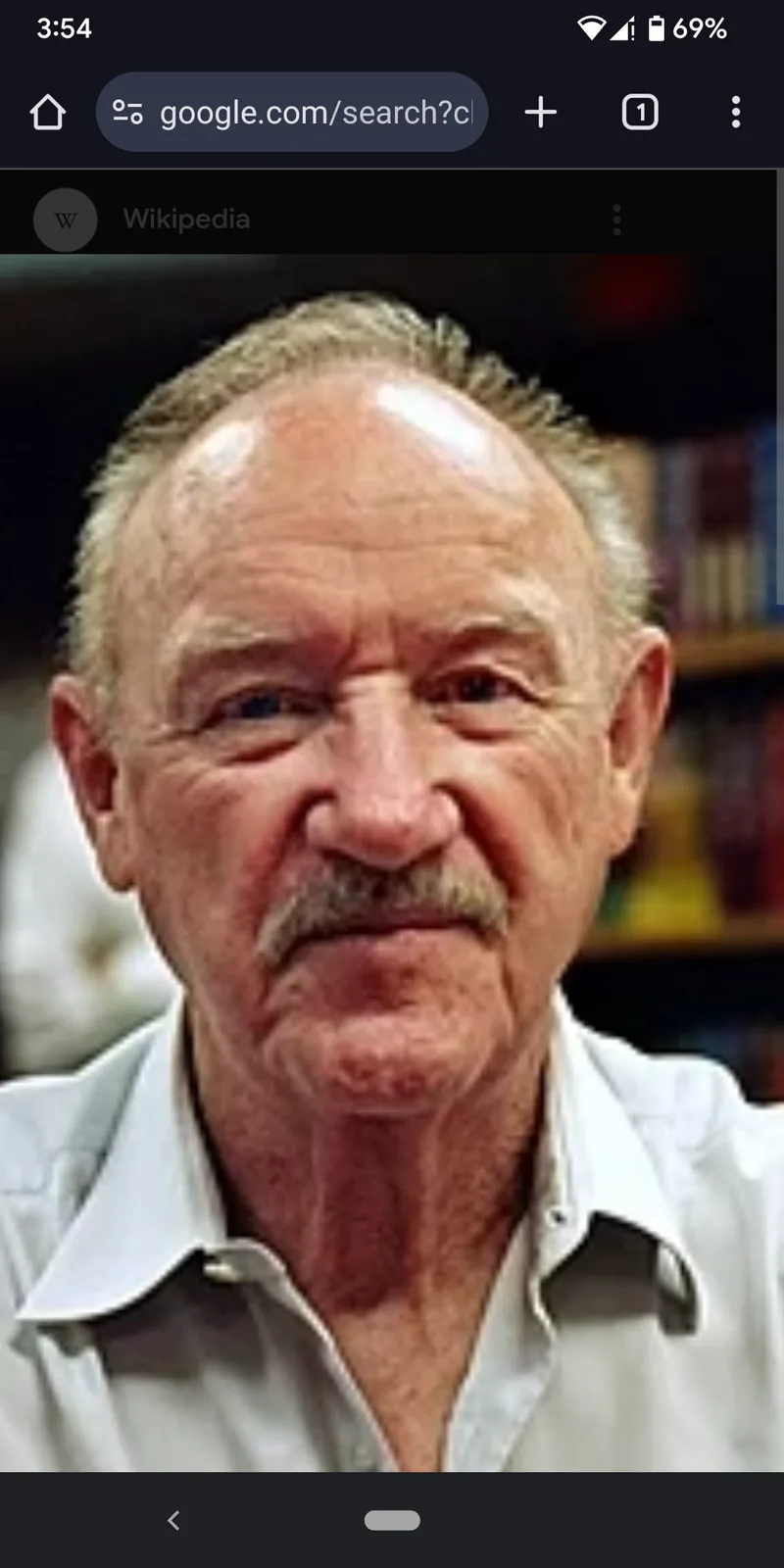 A close-up photo of an older man with a mustache and receding hairline, wearing a white collared shirt, looking directly at…