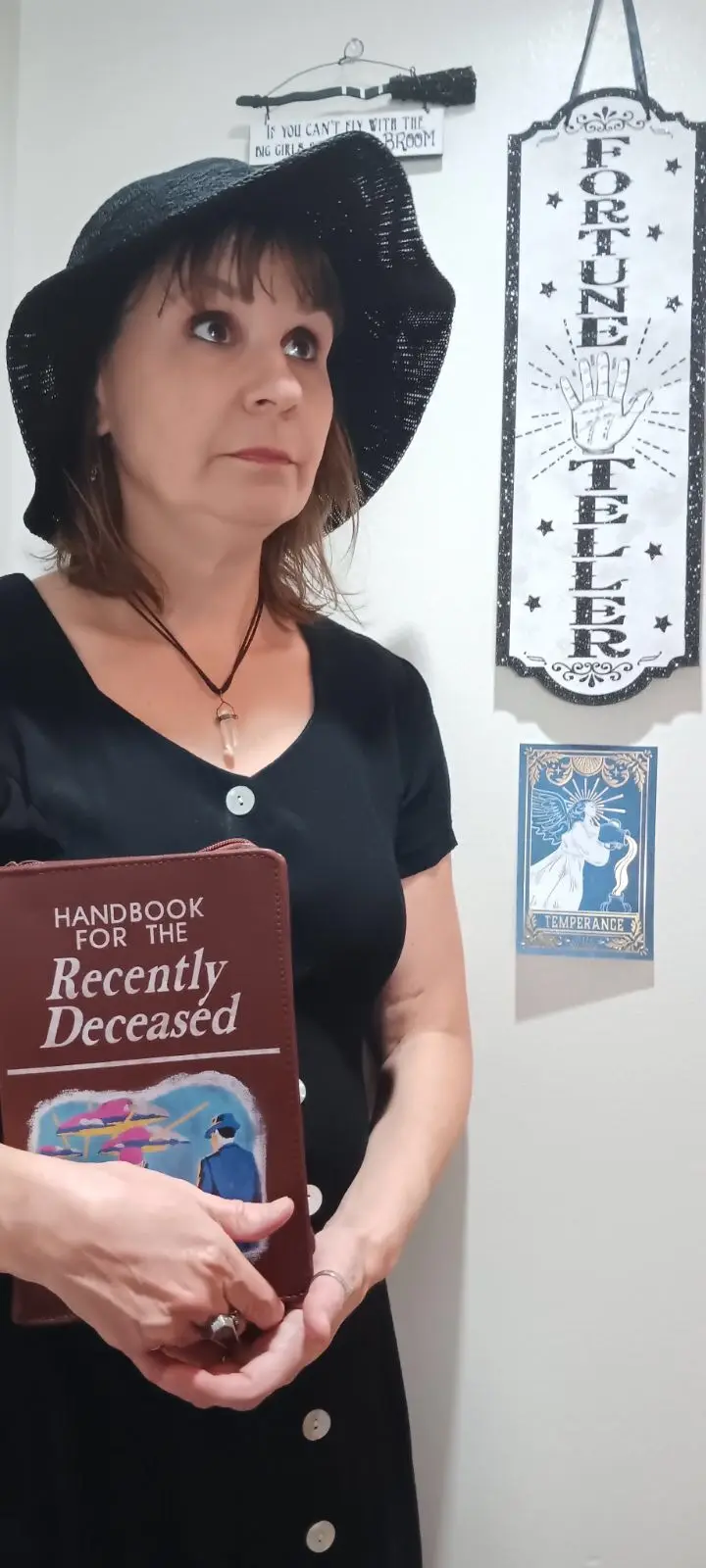 A woman in a black hat and dress holds a brown book titled 'Handbook for the Recently Deceased' while standing in front of a…