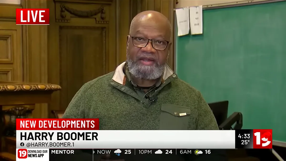 A man with glasses and a gray beard, wearing a green sweater, is shown on a news broadcast. The lower third identifies him…